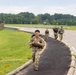 A group of military personnel in camouflage uniforms are conducting a ruck march along a paved path beside a road on what appears to be a military installation. The lead soldier, a smiling young man of apparent Asian descent, is carrying a large rucksack and appears in good spirits. Three additional soldiers follow behind him in a loose formation.