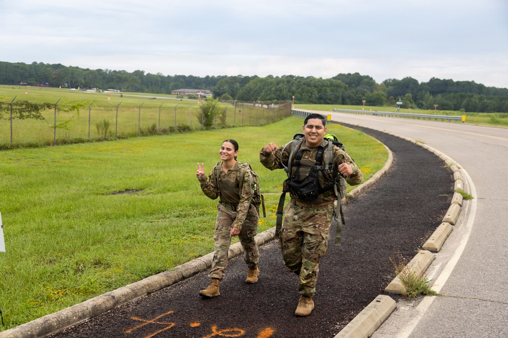 Two soldiers in U.S. Army camouflage (OCP) uniforms are conducting a ruck march along a paved path on what appears to be a military installation. Both are wearing tactical gear and carrying large rucksacks, and they appear to be in good spirits, smiling and gesturing toward the camera. The setting includes open grassy fields, a perimeter fence, and a road consistent with a military base environment.