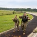 Two soldiers in U.S. Army camouflage (OCP) uniforms are conducting a ruck march along a paved path on what appears to be a military installation. Both are wearing tactical gear and carrying large rucksacks, and they appear to be in good spirits, smiling and gesturing toward the camera. The setting includes open grassy fields, a perimeter fence, and a road consistent with a military base environment.