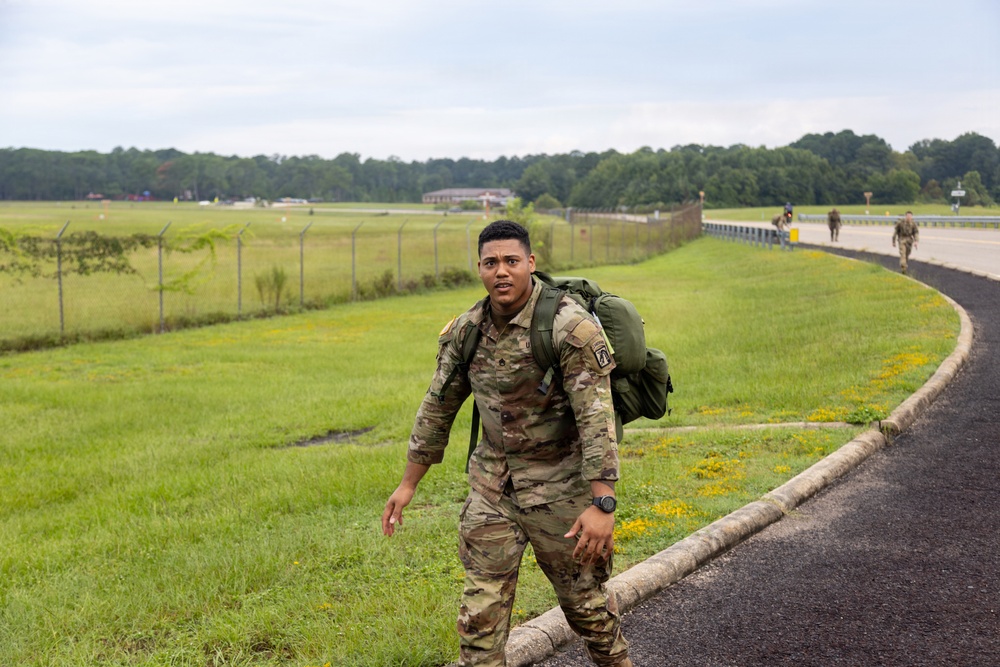 A soldier in U.S. Army OCP (Operational Camouflage Pattern) uniform is conducting a ruck march along a paved path adjacent to a grassy field and road. He is carrying a large green military rucksack and wearing a visible unit patch on his left shoulder. Additional soldiers can be seen in the background performing the same activity.