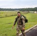 A soldier in U.S. Army OCP (Operational Camouflage Pattern) uniform is conducting a ruck march along a paved path adjacent to a grassy field and road. He is carrying a large green military rucksack and wearing a visible unit patch on his left shoulder. Additional soldiers can be seen in the background performing the same activity.