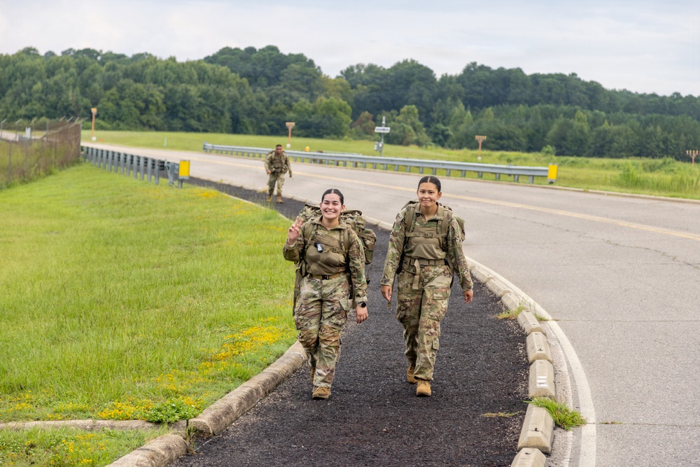 Two female soldiers in U.S. Army OCP (Operational Camouflage Pattern) uniforms walk along a paved pathway beside a road, carrying rucksacks in what appears to be a ruck march or road march exercise. One soldier is smiling and making a peace sign gesture toward the camera. A third soldier in similar uniform is visible further back on the path.