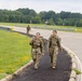 Two female soldiers in U.S. Army OCP (Operational Camouflage Pattern) uniforms walk along a paved pathway beside a road, carrying rucksacks in what appears to be a ruck march or road march exercise. One soldier is smiling and making a peace sign gesture toward the camera. A third soldier in similar uniform is visible further back on the path.
