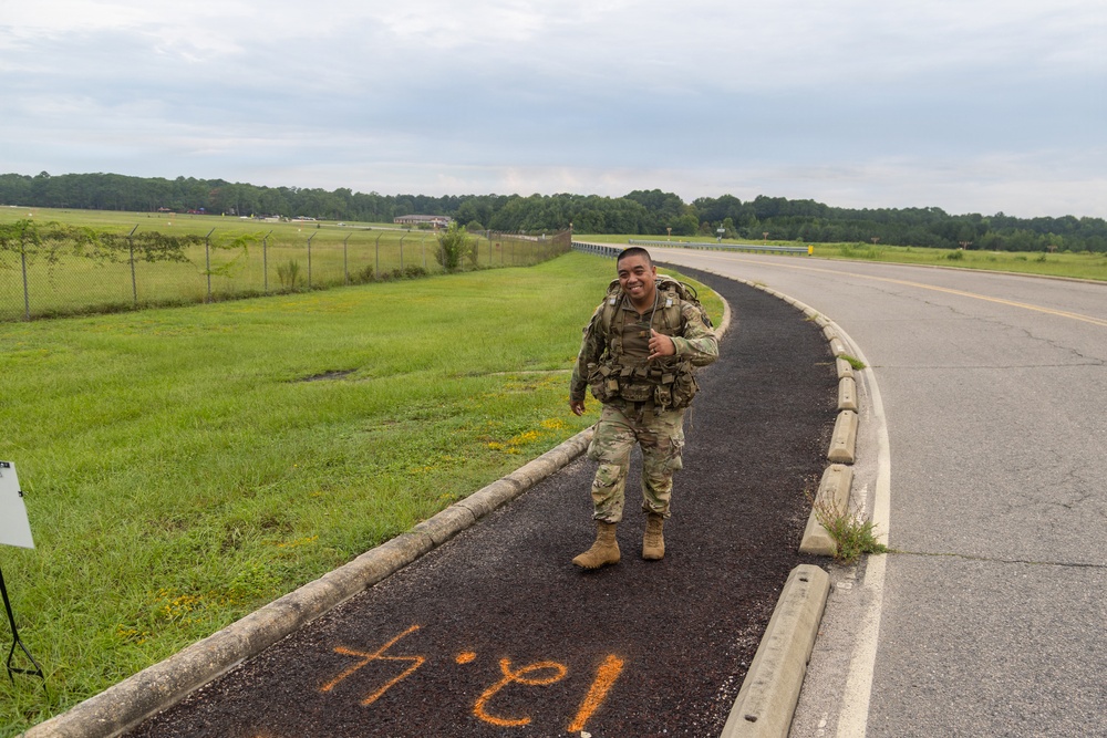 A smiling soldier in full combat uniform and tactical gear walks along a paved path or sidewalk adjacent to a road, giving a thumbs-up gesture. The setting appears to be a military installation with open grassy fields, a perimeter fence, and tree lines in the background. Orange spray-painted markings are visible on the pavement, suggesting a ruck march or physical training event.