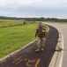 A smiling soldier in full combat uniform and tactical gear walks along a paved path or sidewalk adjacent to a road, giving a thumbs-up gesture. The setting appears to be a military installation with open grassy fields, a perimeter fence, and tree lines in the background. Orange spray-painted markings are visible on the pavement, suggesting a ruck march or physical training event.