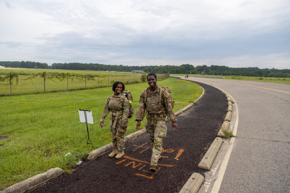 Two soldiers in military camouflage uniforms (OCP/MultiCam pattern) are walking along a paved path, smiling, while carrying rucksacks. Orange spray-painted text reading 'FINISH' (viewed in reverse) is visible on the path beneath them, indicating they are near or at the end of a ruck march or similar military fitness event. Additional participants are visible in the background along the same route.