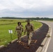Two soldiers in military camouflage uniforms (OCP/MultiCam pattern) are walking along a paved path, smiling, while carrying rucksacks. Orange spray-painted text reading 'FINISH' (viewed in reverse) is visible on the path beneath them, indicating they are near or at the end of a ruck march or similar military fitness event. Additional participants are visible in the background along the same route.