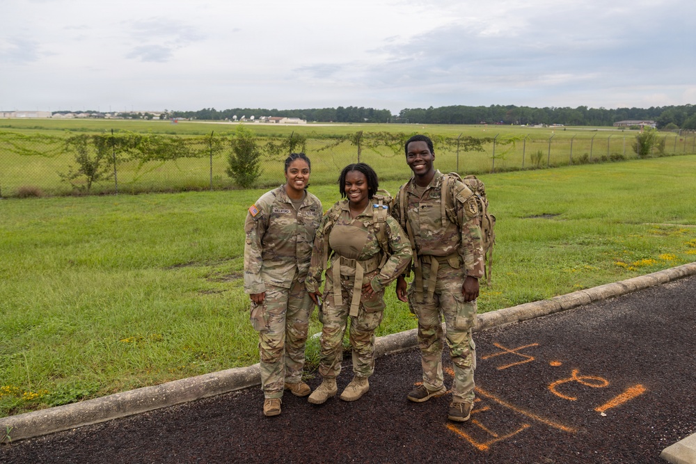 Three U.S. Army soldiers in MultiCam (OCP) camouflage uniforms are posing together and smiling on a paved surface near a grassy field. They are wearing full combat gear including rucksacks and tactical equipment, suggesting they have completed or are participating in a ruck march or field training exercise. The setting appears to be a military installation with open fields and a tree line visible in the background.