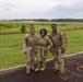 Three U.S. Army soldiers in MultiCam (OCP) camouflage uniforms are posing together and smiling on a paved surface near a grassy field. They are wearing full combat gear including rucksacks and tactical equipment, suggesting they have completed or are participating in a ruck march or field training exercise. The setting appears to be a military installation with open fields and a tree line visible in the background.