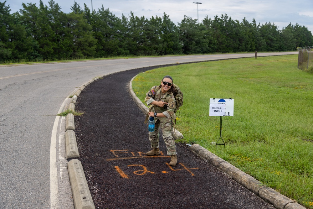 A soldier in full camouflage uniform and tactical gear poses triumphantly at the finish line of a 12.4-mile ruck march or military fitness event. She is pointing down at orange spray-painted text on a dark asphalt path that reads 'FINISH 12.4', with a matching sign on the grass beside her. The setting appears to be a military installation, indicated by the fencing, uniform, and event signage.