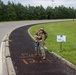 A soldier in full camouflage uniform and tactical gear poses triumphantly at the finish line of a 12.4-mile ruck march or military fitness event. She is pointing down at orange spray-painted text on a dark asphalt path that reads 'FINISH 12.4', with a matching sign on the grass beside her. The setting appears to be a military installation, indicated by the fencing, uniform, and event signage.