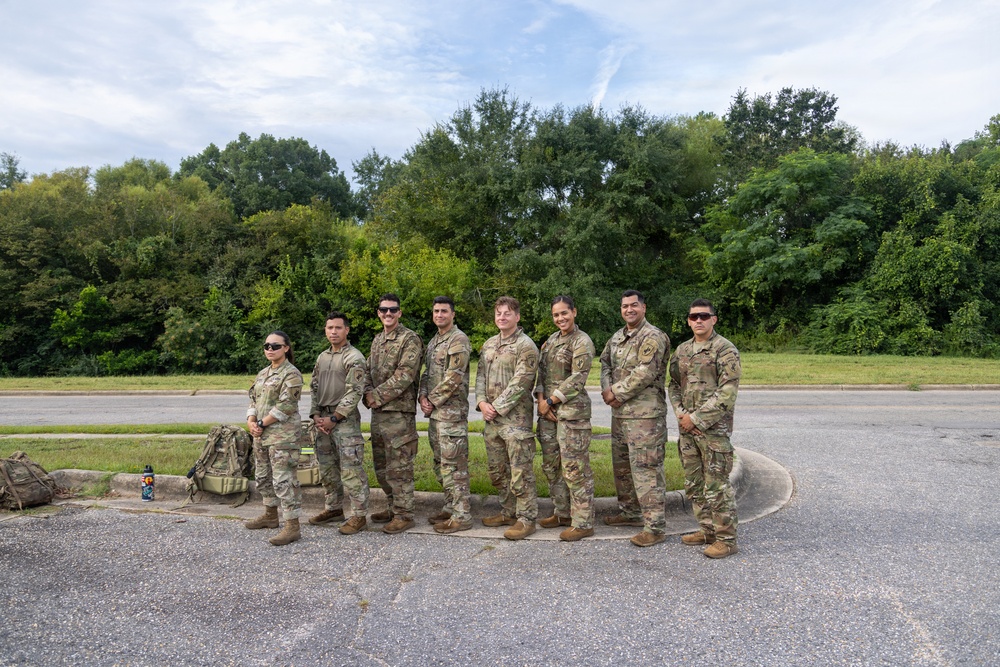A group of eight military personnel in matching camouflage uniforms (OCP/MultiCam pattern) are posed together for a group photo outdoors. They are standing in a line on a paved surface, appearing relaxed and smiling. The setting appears to be a military installation or base area during daytime with lush green trees in the background.