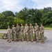 A group of eight military personnel in matching camouflage uniforms (OCP/MultiCam pattern) are posed together for a group photo outdoors. They are standing in a line on a paved surface, appearing relaxed and smiling. The setting appears to be a military installation or base area during daytime with lush green trees in the background.