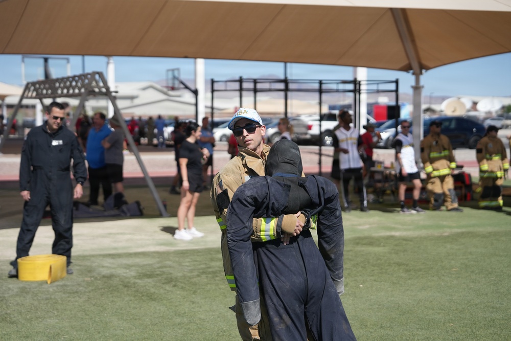 A firefighter in partial turnout gear and a baseball cap is carrying or dragging a weighted rescue dummy across an artificial turf field. The scene appears to be a firefighter fitness competition or training exercise, with multiple participants and spectators visible in the background under shade structures. Additional firefighters in full gear can be seen on the right side of the image.