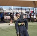 A firefighter in partial turnout gear and a baseball cap is carrying or dragging a weighted rescue dummy across an artificial turf field. The scene appears to be a firefighter fitness competition or training exercise, with multiple participants and spectators visible in the background under shade structures. Additional firefighters in full gear can be seen on the right side of the image.