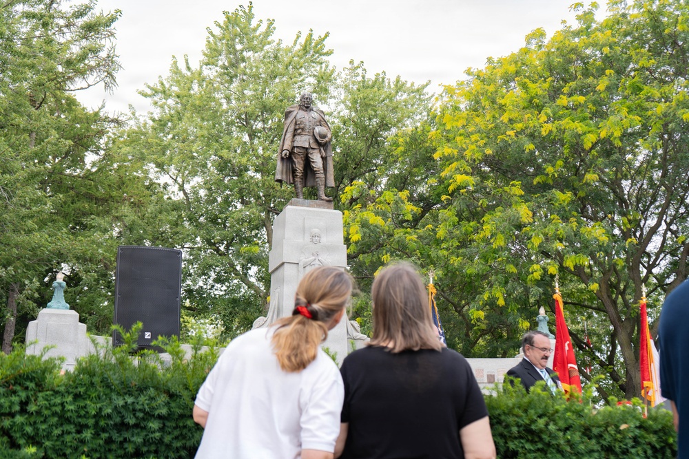 A public memorial or commemorative ceremony is taking place at an outdoor monument featuring a bronze statue of a military figure atop a stone pedestal. Two women are visible from behind observing the event, while a man in a suit stands near military flags on the right side. The setting includes lush green trees and appears to be a park or public square.