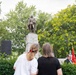A public memorial or commemorative ceremony is taking place at an outdoor monument featuring a bronze statue of a military figure atop a stone pedestal. Two women are visible from behind observing the event, while a man in a suit stands near military flags on the right side. The setting includes lush green trees and appears to be a park or public square.