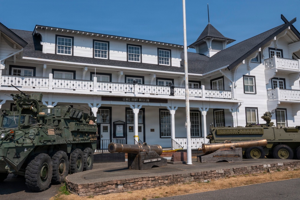 The image shows the Lewis Army Museum, a two-story white colonial-style building with black trim, located on a military installation. In front of the building, several military vehicles and historical artillery pieces are displayed as outdoor exhibits. A flagpole stands prominently in the foreground, and a sign reading 'Lewis Army Museum' is visible above the entrance.