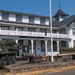 The image shows the Lewis Army Museum, a two-story white colonial-style building with black trim, located on a military installation. In front of the building, several military vehicles and historical artillery pieces are displayed as outdoor exhibits. A flagpole stands prominently in the foreground, and a sign reading 'Lewis Army Museum' is visible above the entrance.