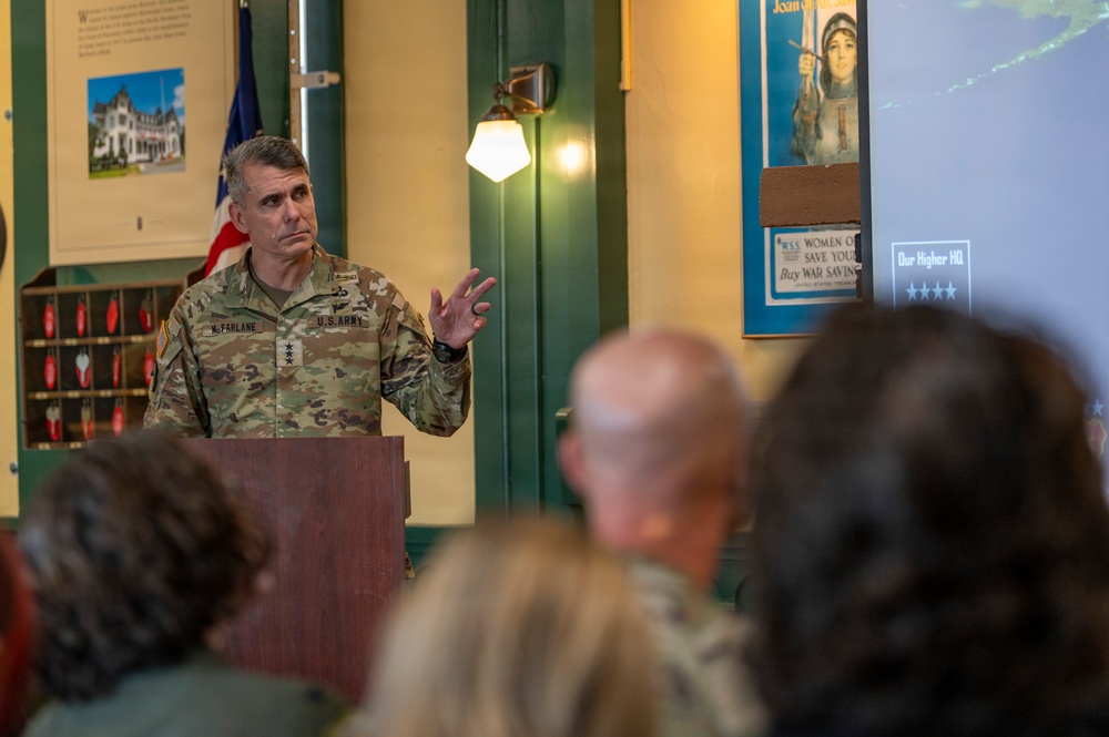 A U.S. Army officer with the name tag 'McFARLANE' and three-star general rank insignia is speaking at a wooden podium before an audience. The setting appears to be a museum or historical exhibit space, with vintage WWI-era war bond posters visible on the wall behind him. He is gesturing with his right hand while addressing the seated crowd.
