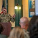 A U.S. Army officer with the name tag 'McFARLANE' and three-star general rank insignia is speaking at a wooden podium before an audience. The setting appears to be a museum or historical exhibit space, with vintage WWI-era war bond posters visible on the wall behind him. He is gesturing with his right hand while addressing the seated crowd.