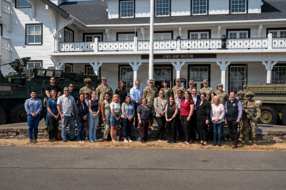 A large group of approximately 30 people, including both military personnel in camouflage uniforms and civilians in business casual attire, pose for a group photo in front of the Lewis Army Museum. The historic white two-story building serves as the backdrop, flanked by military vehicles including what appear to be Stryker armored vehicles. The setting suggests an official visit, community engagement event, or employer support program at Joint Base Lewis-McChord.