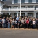 A large group of approximately 30 people, including both military personnel in camouflage uniforms and civilians in business casual attire, pose for a group photo in front of the Lewis Army Museum. The historic white two-story building serves as the backdrop, flanked by military vehicles including what appear to be Stryker armored vehicles. The setting suggests an official visit, community engagement event, or employer support program at Joint Base Lewis-McChord.