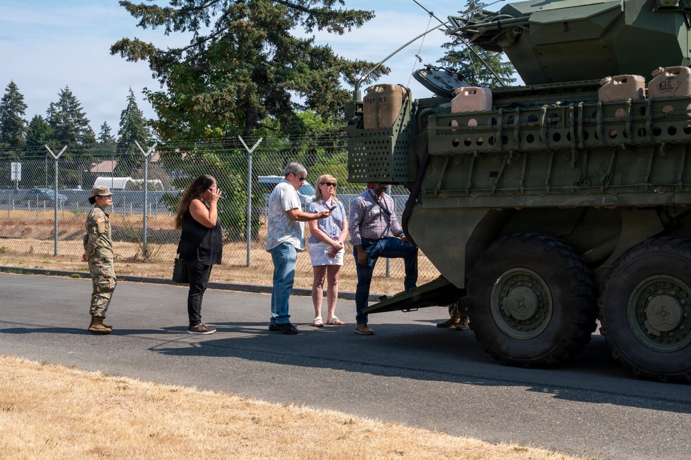 A group of civilians is being briefed or given a tour near a large military armored vehicle, likely a Stryker, on what appears to be a military installation. One uniformed soldier in camouflage (OCP pattern) is present, appearing to guide or escort the civilian visitors. The setting is outdoors on a paved road with a chain-link perimeter fence and trees visible in the background.