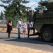 A group of civilians is being briefed or given a tour near a large military armored vehicle, likely a Stryker, on what appears to be a military installation. One uniformed soldier in camouflage (OCP pattern) is present, appearing to guide or escort the civilian visitors. The setting is outdoors on a paved road with a chain-link perimeter fence and trees visible in the background.