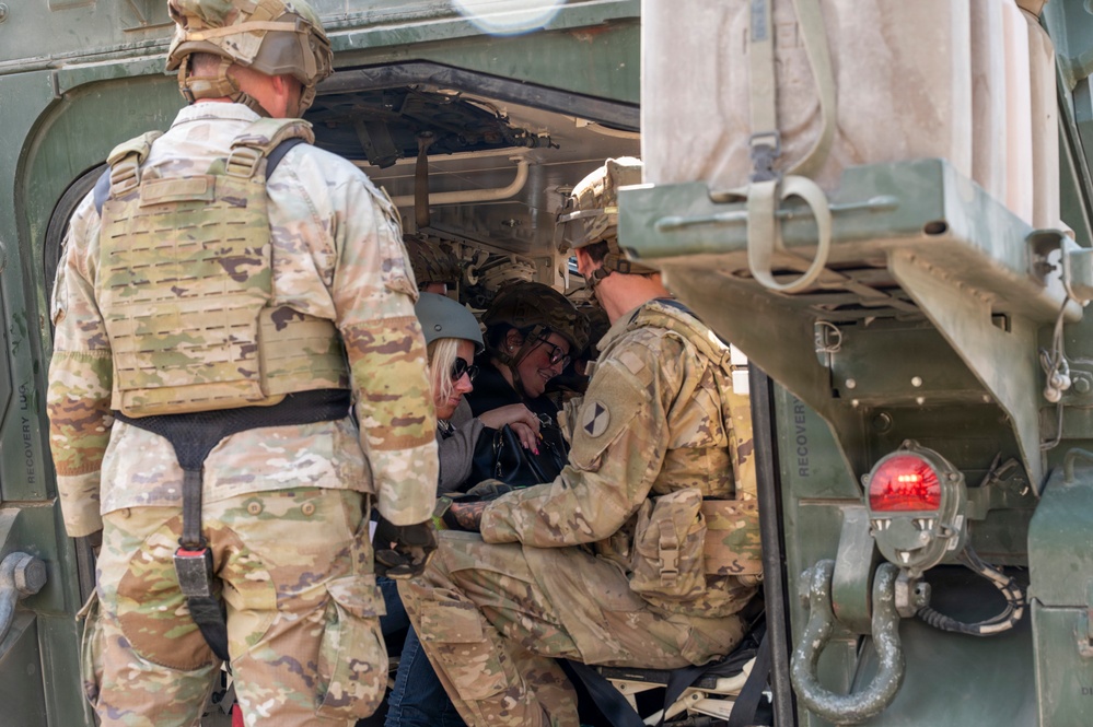 The image shows U.S. Army soldiers in OCP (Operational Camouflage Pattern) uniforms interacting with two civilian women inside or near a military vehicle, likely a HMMWV (Humvee). The soldiers appear to be giving the civilians a demonstration or familiarization tour of the vehicle's interior. A unit patch consistent with the 1st Cavalry Division is visible on one soldier's sleeve.