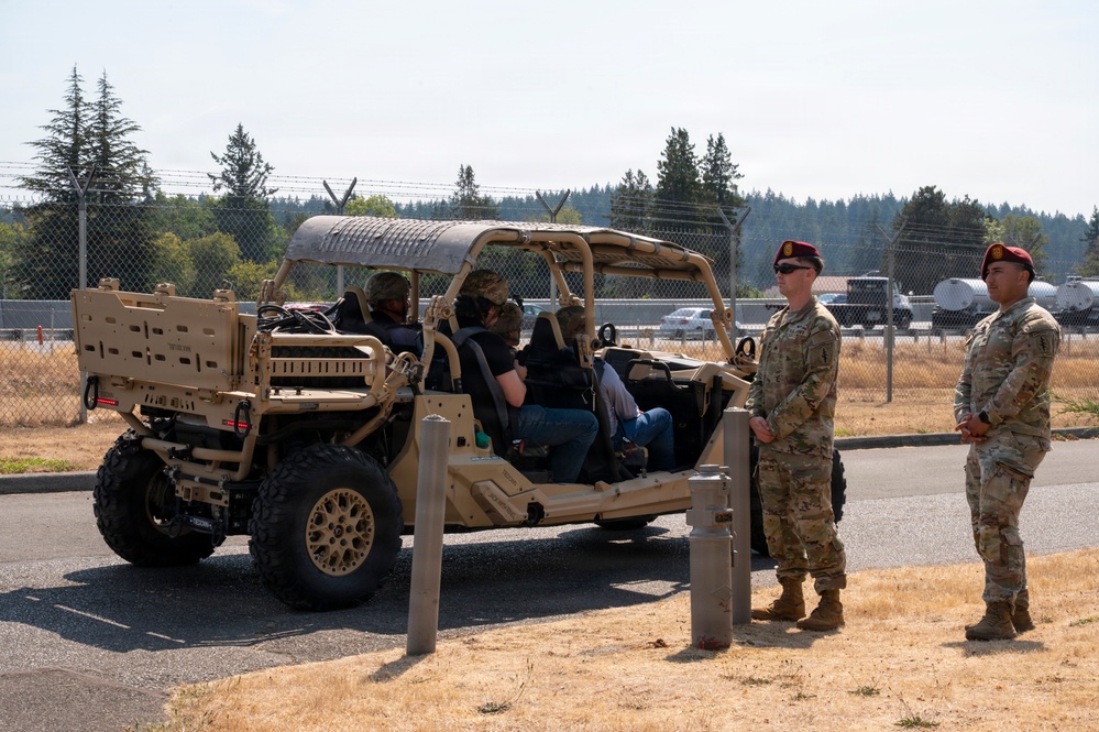 Two uniformed military personnel wearing maroon/red berets and MultiCam (OCP) uniforms stand beside a tan military utility terrain vehicle (UTV), likely a Polaris MRZR or similar tactical vehicle. Two civilians are seated inside the vehicle, suggesting a demonstration, briefing, or familiarization event. The setting appears to be a military installation with a perimeter fence, open field, and tree line visible in the background.