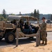 Two uniformed military personnel wearing maroon/red berets and MultiCam (OCP) uniforms stand beside a tan military utility terrain vehicle (UTV), likely a Polaris MRZR or similar tactical vehicle. Two civilians are seated inside the vehicle, suggesting a demonstration, briefing, or familiarization event. The setting appears to be a military installation with a perimeter fence, open field, and tree line visible in the background.