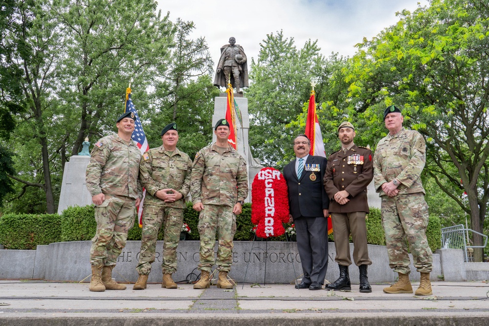 Six individuals pose in front of a memorial monument during what appears to be a wreath-laying ceremony. Four are wearing US Army multicam uniforms, one is in a brown military dress uniform with medals, and one is in civilian attire with a suit and tie. A large red floral wreath with white lettering reading 'USA' is prominently displayed in front of the monument's base.