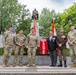 Six individuals pose in front of a memorial monument during what appears to be a wreath-laying ceremony. Four are wearing US Army multicam uniforms, one is in a brown military dress uniform with medals, and one is in civilian attire with a suit and tie. A large red floral wreath with white lettering reading 'USA' is prominently displayed in front of the monument's base.
