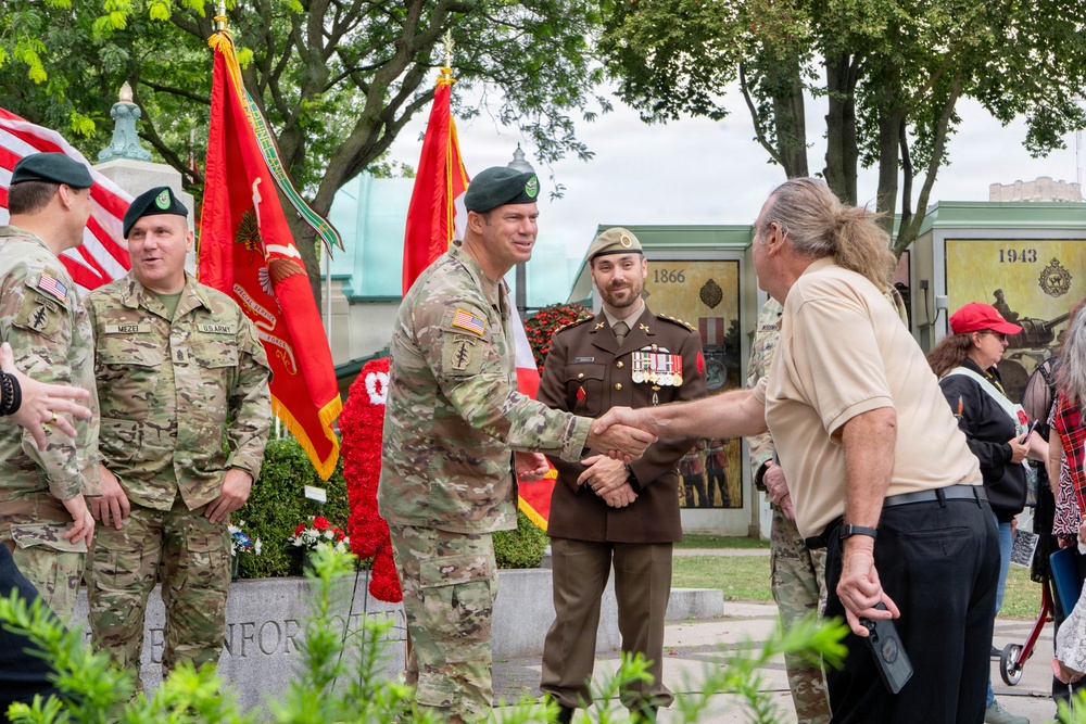 A military ceremony taking place outdoors at what appears to be a memorial or monument site. U.S. Army Special Forces soldiers wearing green berets are greeting civilians and a foreign military officer, with ceremonial flags including the U.S. flag and red military unit colors visible in the background. The setting includes memorial plaques with years '1866' and '1943' visible.
