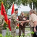 A military ceremony taking place outdoors at what appears to be a memorial or monument site. U.S. Army Special Forces soldiers wearing green berets are greeting civilians and a foreign military officer, with ceremonial flags including the U.S. flag and red military unit colors visible in the background. The setting includes memorial plaques with years '1866' and '1943' visible.