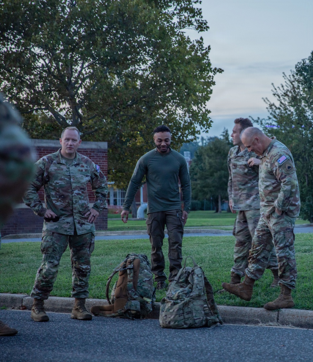 A group of military personnel in camouflage uniforms are gathered outdoors on what appears to be a military installation at dusk or dawn. Several tactical backpacks/rucksacks are placed on the ground near the curb, suggesting preparation for or completion of a ruck march or field exercise. The setting includes a brick building, manicured grass, and mature trees in the background.