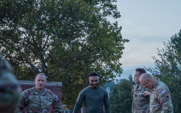 A group of military personnel in camouflage uniforms are gathered outdoors on what appears to be a military installation at dusk or dawn. Several tactical backpacks/rucksacks are placed on the ground near the curb, suggesting preparation for or completion of a ruck march or field exercise. The setting includes a brick building, manicured grass, and mature trees in the background.