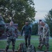 A group of military personnel in camouflage uniforms are gathered outdoors on what appears to be a military installation at dusk or dawn. Several tactical backpacks/rucksacks are placed on the ground near the curb, suggesting preparation for or completion of a ruck march or field exercise. The setting includes a brick building, manicured grass, and mature trees in the background.