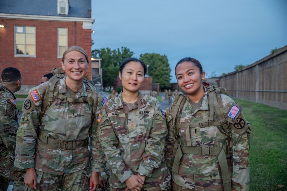 Three female U.S. Army soldiers in Operational Camouflage Pattern (OCP) uniforms pose together outdoors, smiling at the camera. They are wearing tactical gear including rucksacks and appear to be at a military installation, likely during or after a training event. The setting appears to be early morning or evening based on the ambient light.