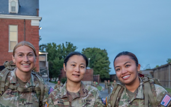 Three female U.S. Army soldiers in Operational Camouflage Pattern (OCP) uniforms pose together outdoors, smiling at the camera. They are wearing tactical gear including rucksacks and appear to be at a military installation, likely during or after a training event. The setting appears to be early morning or evening based on the ambient light.