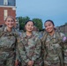 Three female U.S. Army soldiers in Operational Camouflage Pattern (OCP) uniforms pose together outdoors, smiling at the camera. They are wearing tactical gear including rucksacks and appear to be at a military installation, likely during or after a training event. The setting appears to be early morning or evening based on the ambient light.
