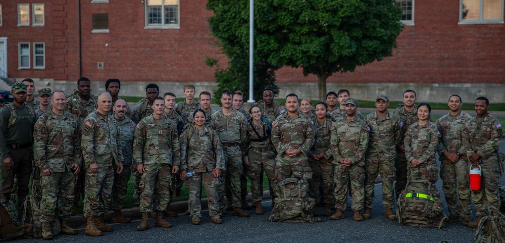 A large group of approximately 30 military personnel in matching camouflage uniforms (OCP/MultiCam pattern) pose together for a group photograph outdoors. They are standing in front of a red brick building on what appears to be a military installation or base. The group includes both male and female soldiers, and some individuals have tactical backpacks and gear visible.