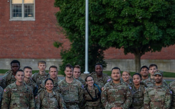 A large group of approximately 30 military personnel in matching camouflage uniforms (OCP/MultiCam pattern) pose together for a group photograph outdoors. They are standing in front of a red brick building on what appears to be a military installation or base. The group includes both male and female soldiers, and some individuals have tactical backpacks and gear visible.