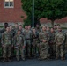 A large group of approximately 30 military personnel in matching camouflage uniforms (OCP/MultiCam pattern) pose together for a group photograph outdoors. They are standing in front of a red brick building on what appears to be a military installation or base. The group includes both male and female soldiers, and some individuals have tactical backpacks and gear visible.