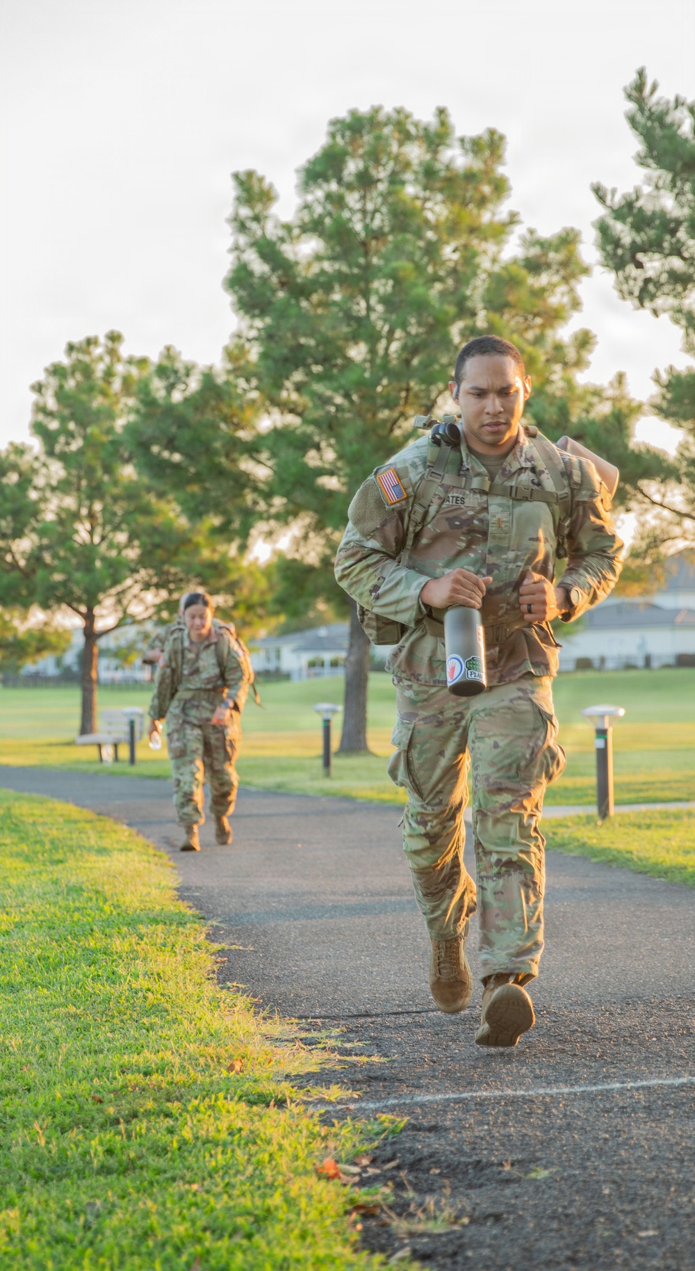 Two U.S. Army soldiers in Operational Camouflage Pattern (OCP) uniforms are conducting a ruck march or physical training exercise along a paved path in a well-maintained park or military installation. The lead soldier, whose name tape reads 'ATES,' carries a metal water bottle and wears a backpack, with an American flag patch visible on the right shoulder. A second soldier follows behind on the same path, also carrying a rucksack.