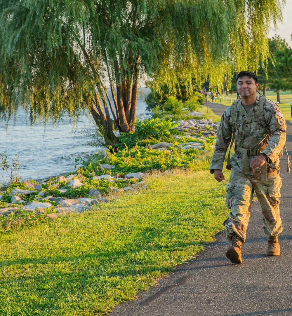 A U.S. Army soldier in Operational Camouflage Pattern (OCP) uniform is walking along a paved path beside a waterfront area. The soldier is wearing a tactical vest/ruck and appears to be smiling, suggesting participation in a ruck march or physical training exercise. The setting features lush green grass, weeping willow trees, and a rocky shoreline along a body of water.