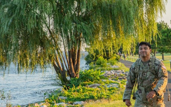 A U.S. Army soldier in Operational Camouflage Pattern (OCP) uniform is walking along a paved path beside a waterfront area. The soldier is wearing a tactical vest/ruck and appears to be smiling, suggesting participation in a ruck march or physical training exercise. The setting features lush green grass, weeping willow trees, and a rocky shoreline along a body of water.