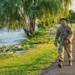 A U.S. Army soldier in Operational Camouflage Pattern (OCP) uniform is walking along a paved path beside a waterfront area. The soldier is wearing a tactical vest/ruck and appears to be smiling, suggesting participation in a ruck march or physical training exercise. The setting features lush green grass, weeping willow trees, and a rocky shoreline along a body of water.