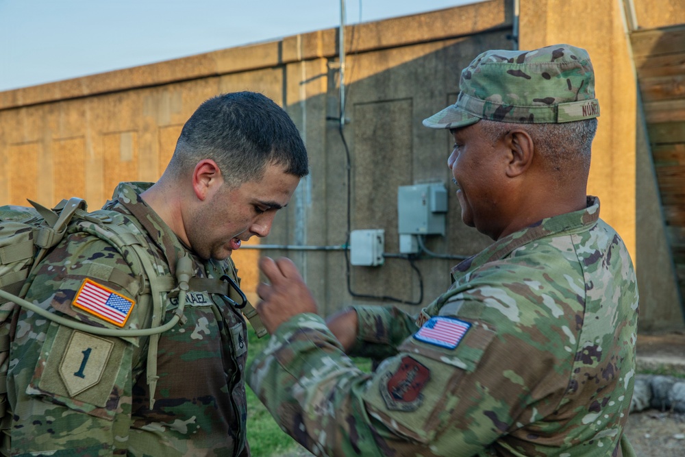 Two U.S. Army soldiers in Operational Camouflage Pattern (OCP) uniforms are engaged in what appears to be an equipment inspection or pinning ceremony outdoors near a concrete structure. The soldier on the left wears a '1st Infantry Division' shoulder patch and a rucksack, while the soldier on the right, whose name tape reads 'NUNEZ', is gesturing toward the other soldier's chest. The interaction suggests a formal or instructional moment between the two service members.